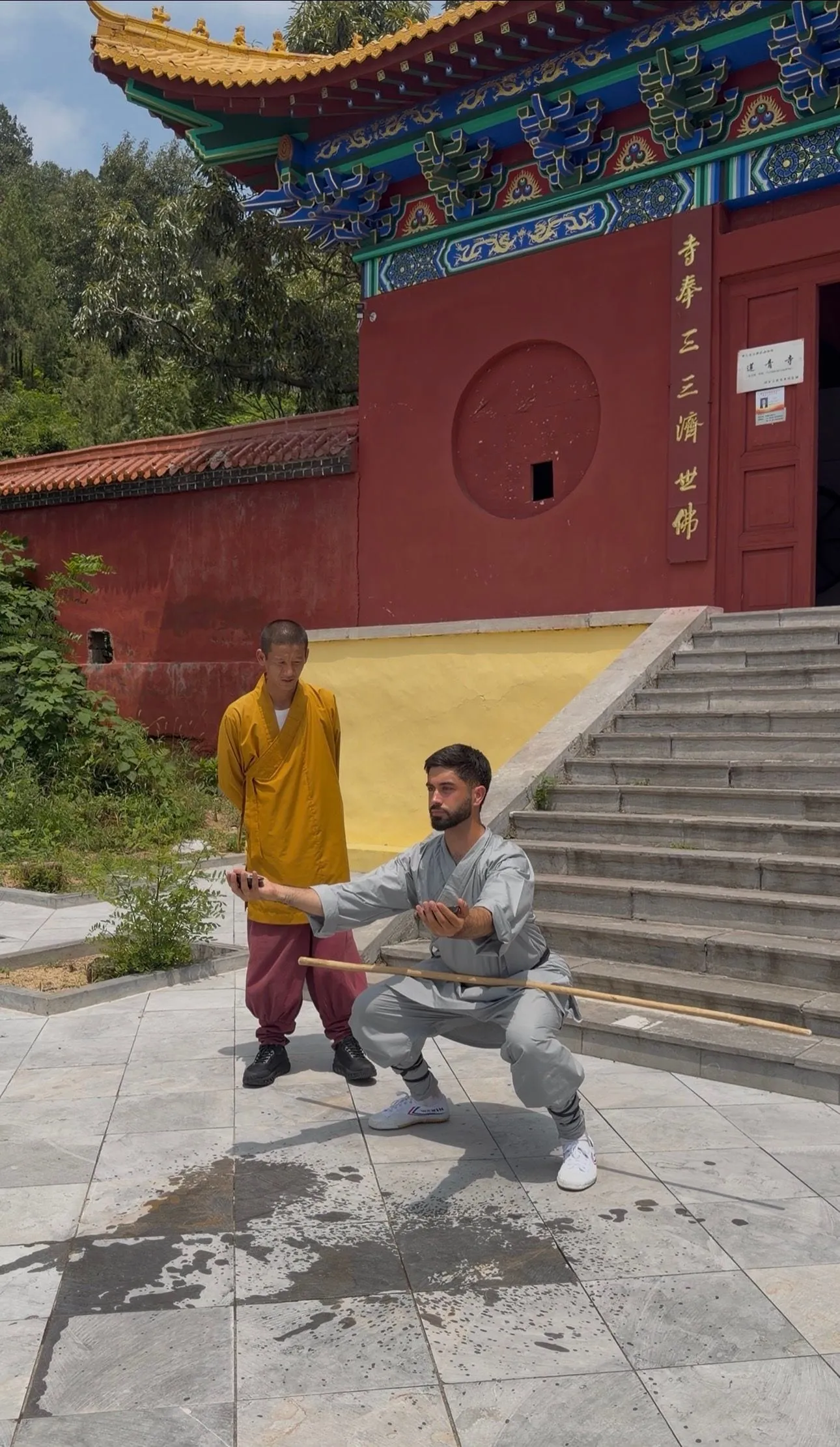 Yassine, en tenue grise, tient un bâton en position basse, guidé par un maître en robe safran devant un temple aux murs rouges.