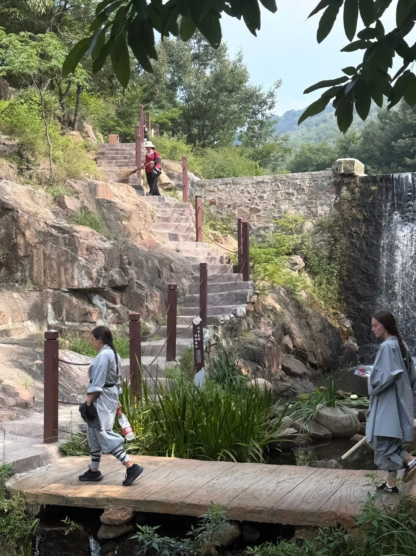 Deux élèves en tenue grise traversent une passerelle en bois au pied d’une cascade.