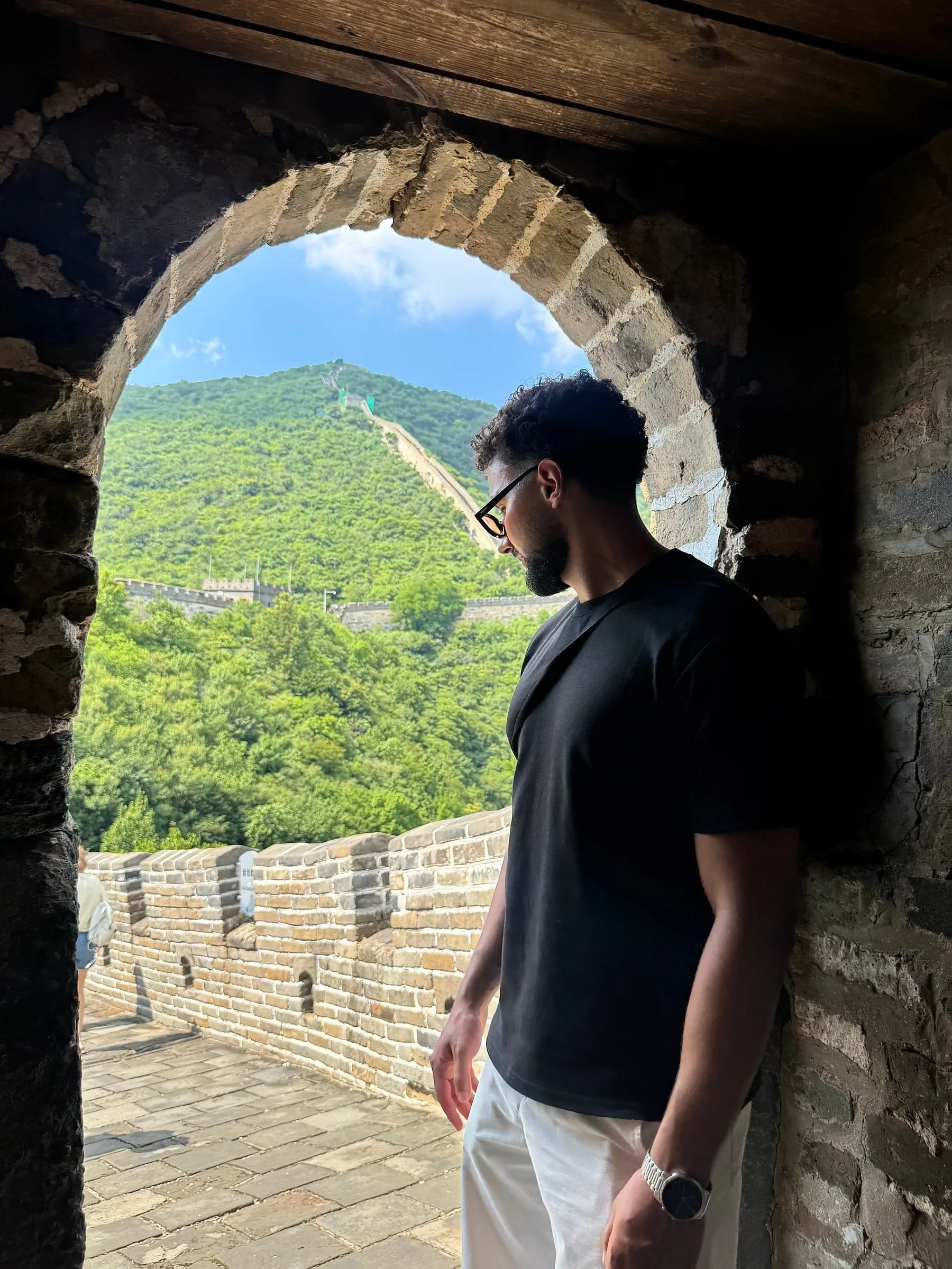 Yassine, en t-shirt noir, adossé à l’encadrement d’une ouverture voûtée en pierre ; derrière lui, la Muraille grimpe une colline boisée.